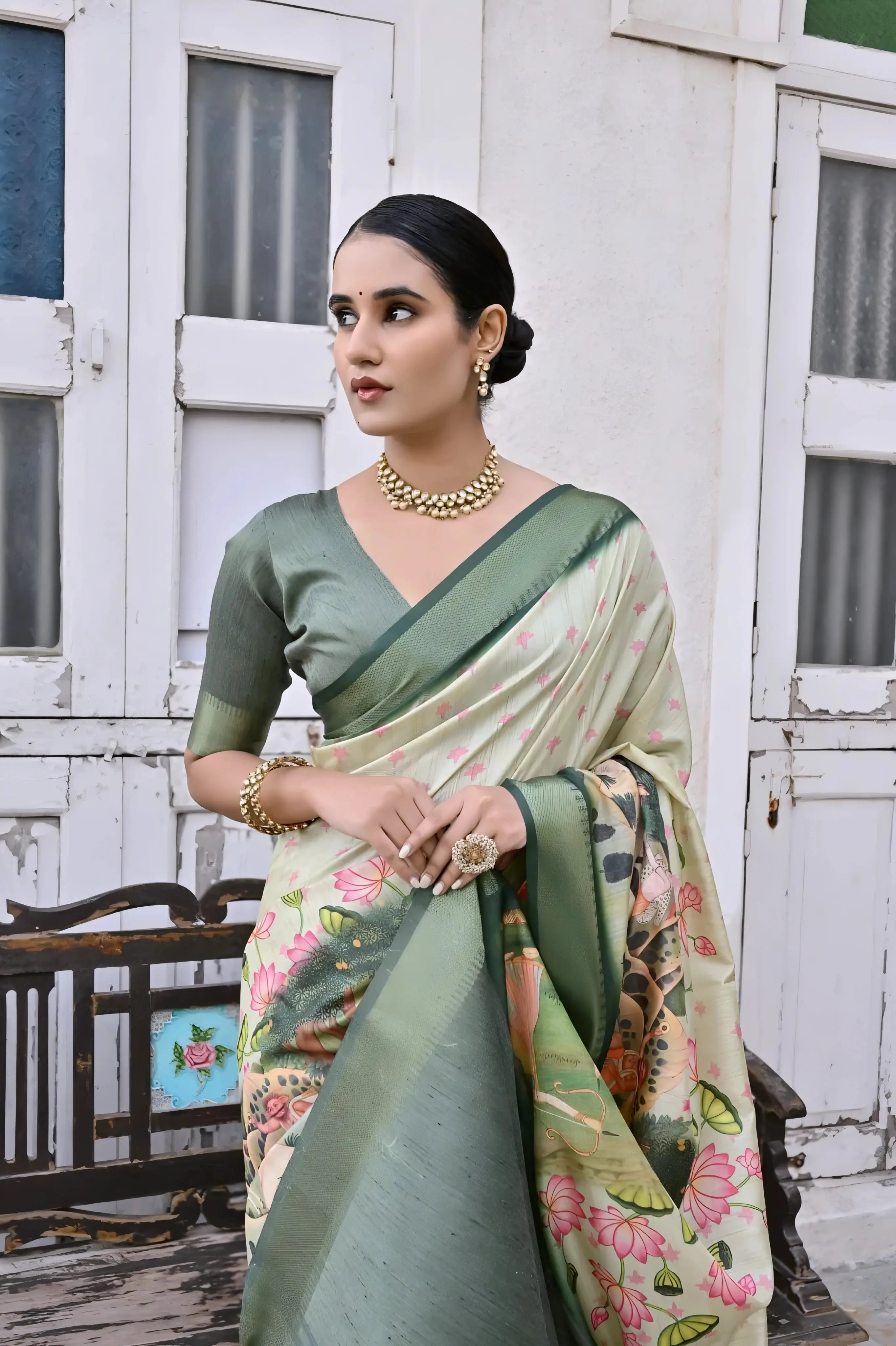 A woman models an elegant Off-White Soft Silk Saree featuring a large-scale, colorful floral and foliage Pichwai-style print. The saree has a wide Dark Olive Green border and pallu. She wears a matching short-sleeve green blouse and a gold Kundan choker necklace, posing outdoors next to antique white doors.